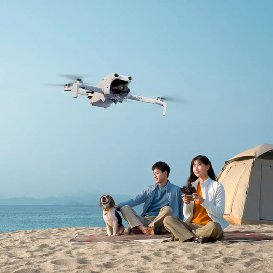 Two people sitting on a beach with a drone flying above them, a tent in the background.